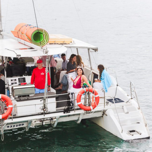 A group of people relax and socialize on a white catamaran boat with orange life rings, floating on calm water. Some passengers are seated, while others stand or pose near the edge, enjoying the outing.