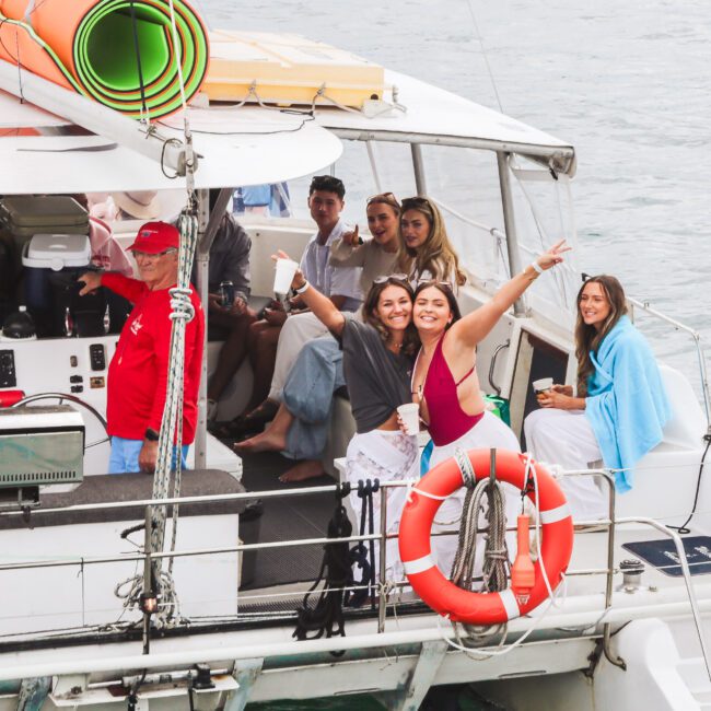 A group of people smile and pose on a boat, some wrapped in towels. Two women in the foreground point and wave at the camera, while others sit or stand behind them. The boat is on calm water, with rolled-up mats on the roof.