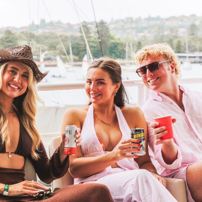 Three people sit on a boat, smiling and holding drinks. One woman wears a brown hat and dress, another wears a white outfit, and a man in pink holds a red cup. Sailboats and trees are visible in the background.