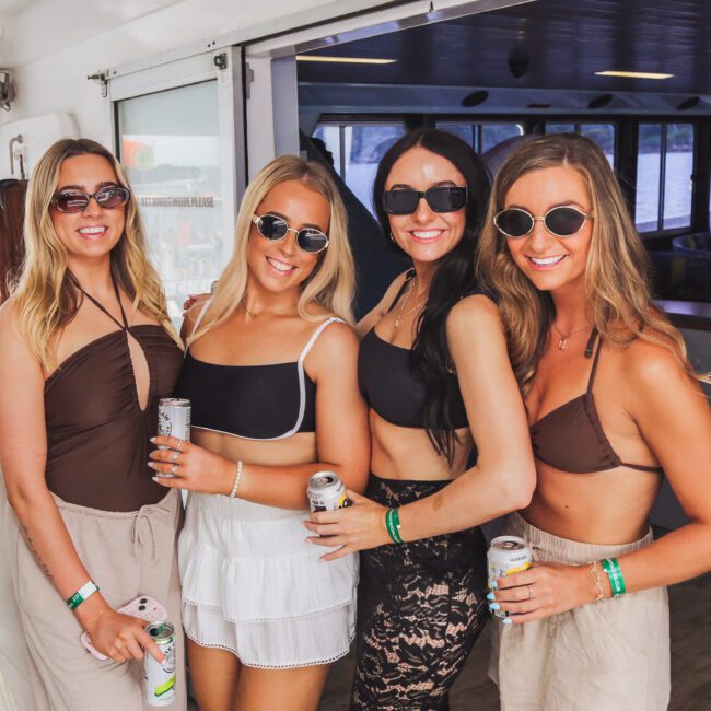 Four women wearing sunglasses and beachwear smile and pose together on a boat, each holding a canned drink. Sunlight streams inside and water can be seen through the windows in the background.