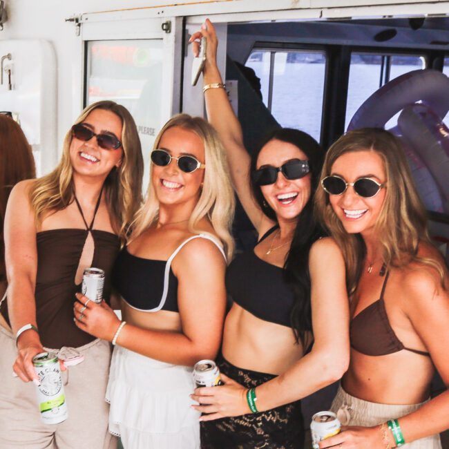 Four women in sunglasses and casual summer outfits smile and pose together on a boat, each holding a canned drink. They appear to be enjoying a sunny day and a fun outing.