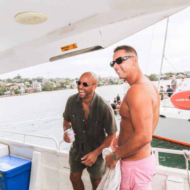 Two men wearing sunglasses smile and stand on a boat, with water and another boat in the background; one man is shirtless, the other wears a striped shirt. The mood is relaxed and cheerful.