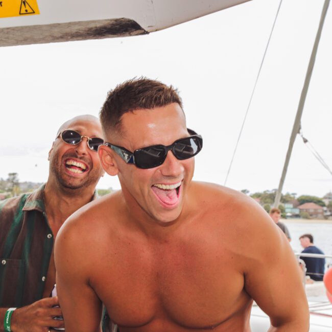 Two men wearing sunglasses are laughing joyfully on a boat. One is shirtless, and the other wears a green shirt. The background shows water, buildings, and a “Mind your head” sign above them.