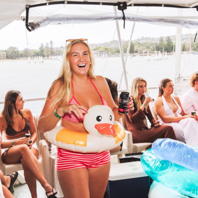 A smiling woman in a red-striped swimsuit and inflatable duck float holds drinks on a boat with other people. The group sits and talks, enjoying a sunny day on the water with trees and buildings visible in the background.