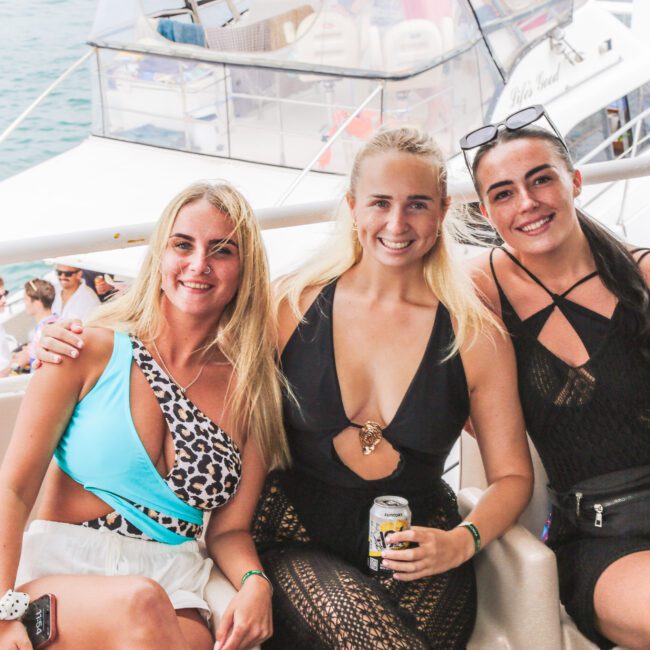 Three smiling women in swimsuits sit close together on a boat, enjoying drinks. Other people and a large white yacht are visible in the background on the water.