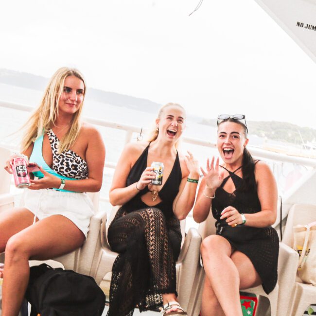 Three women sit on a boat deck, smiling and laughing while holding drinks. One wears white shorts and a patterned top, the others wear black outfits. The background shows water and distant land. A sign reads “NO JUMPING FROM UPPER DECK.”.
