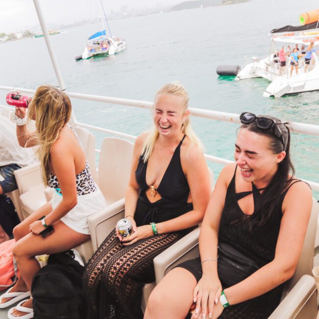 Three women sitting and laughing on a boat with drinks in hand, while another person sits nearby. The boat is on the water with other boats visible in the background on a sunny day.