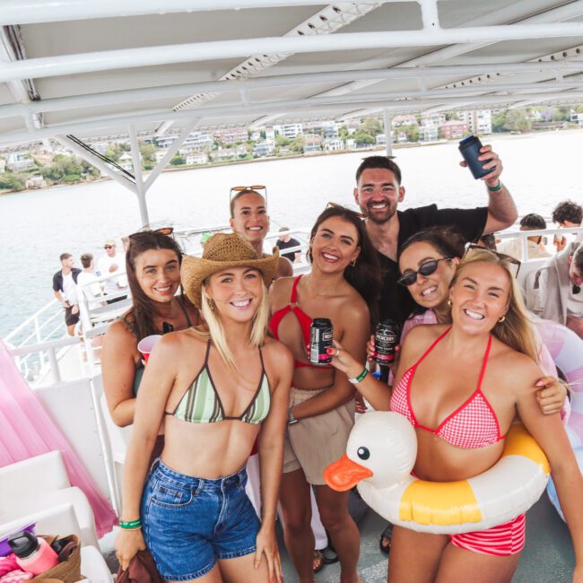 A group of smiling young adults in swimsuits poses for a photo on a boat, holding drinks and inflatable pool floats. The background shows water and a shoreline with buildings under a sunny sky.