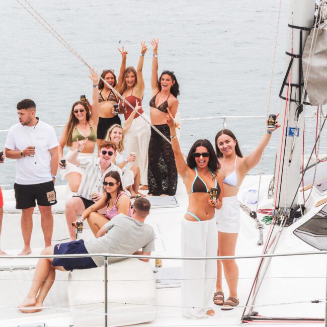 A group of people in swimsuits and casual summer clothes celebrate on a yacht, holding drinks, smiling, and posing for a photo with the ocean in the background.