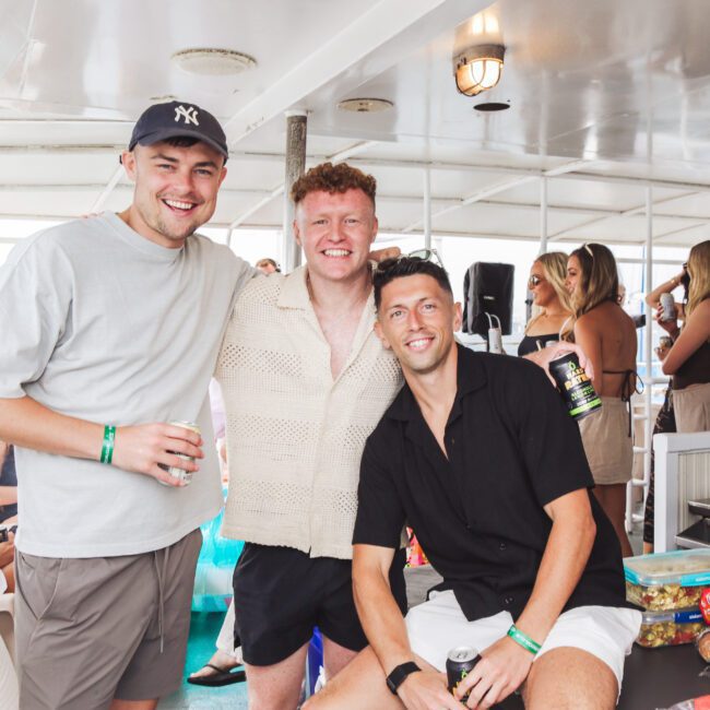 Three young men smile for a photo on a boat, surrounded by people. Two stand while one sits, all casually dressed and wearing wristbands. The background shows snacks and others mingling on the boat.