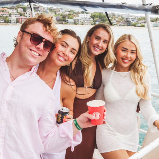 Four young adults, three women and one man, smile and pose together on a boat. The man wears sunglasses and light clothing, while the women wear stylish dresses. The background shows water and a shoreline with houses.