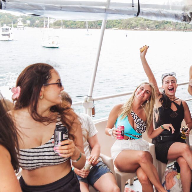 Four women in swimsuits are smiling and celebrating on a boat, holding drinks. The background shows water, other boats, and a green, tree-lined shore. The atmosphere is lively and joyful.