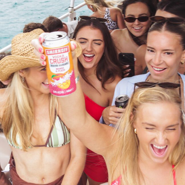 A group of young women in swimsuits smile and celebrate on a boat. One woman holds up a can of Smirnoff Vodka Crush. The background shows more people and the blue water.