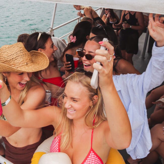 A group of young people in swimsuits and summer clothes dance and celebrate on a boat, holding drinks and smiling, with the ocean visible in the background.