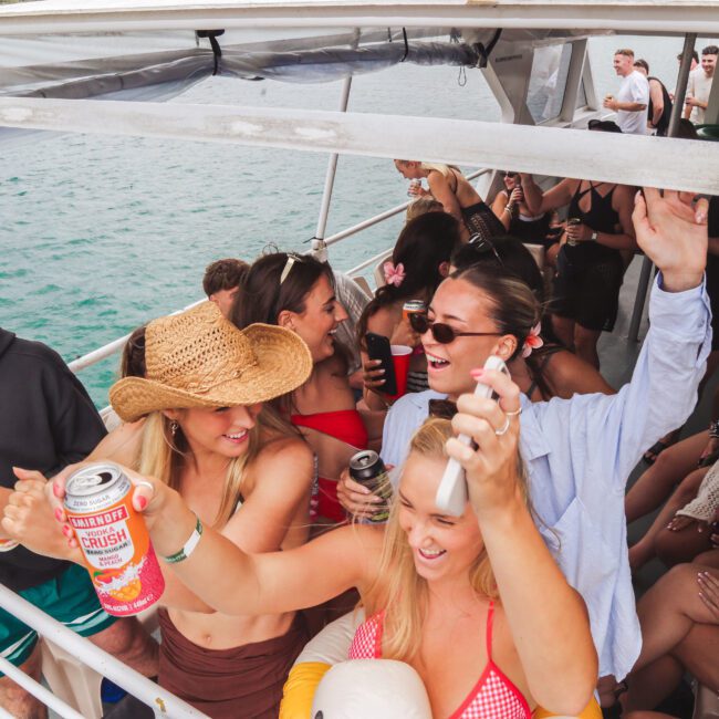 A group of young adults enjoy a lively party on a boat, holding drinks, dancing, and smiling. The boat is on the water, and everyone appears to be having fun in casual summer clothing.
