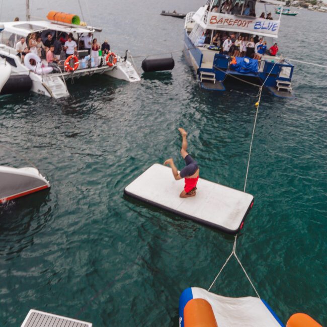 A person is mid-flip on a floating mat in the water between two boats full of people, with other boats and a shoreline visible in the background.