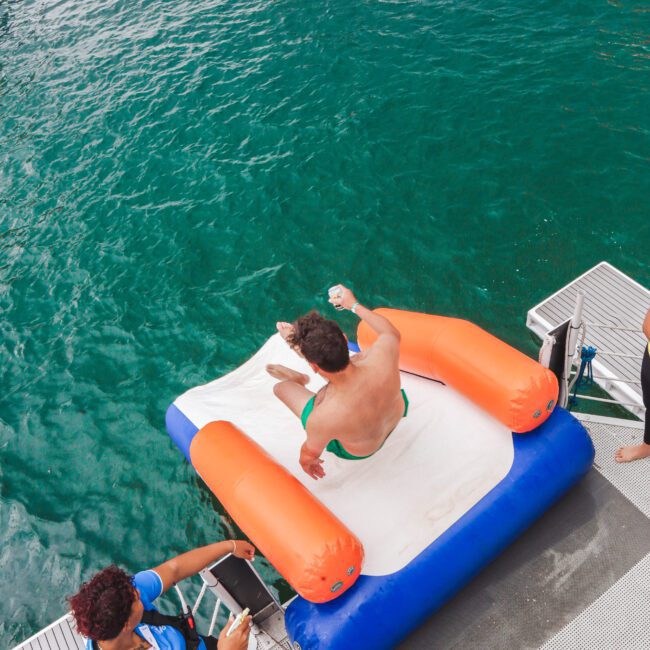 A man in swim trunks sits on an inflatable float at the edge of a dock above turquoise water, while three people in blue shirts stand nearby, watching.