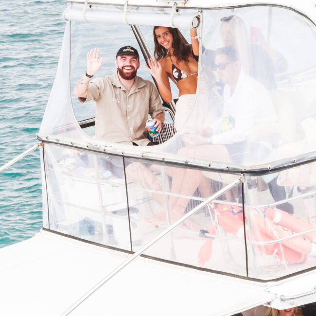 A man and woman on the upper deck of a boat wave at the camera, smiling. Several people are visible on the lower deck, enjoying drinks and socializing near the water.