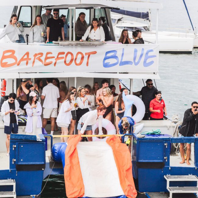 A group of people in summer attire gather on the deck of a blue and white boat named "Barefoot Blue," enjoying a lively party with drinks, inflatables, and music, surrounded by water and other boats.