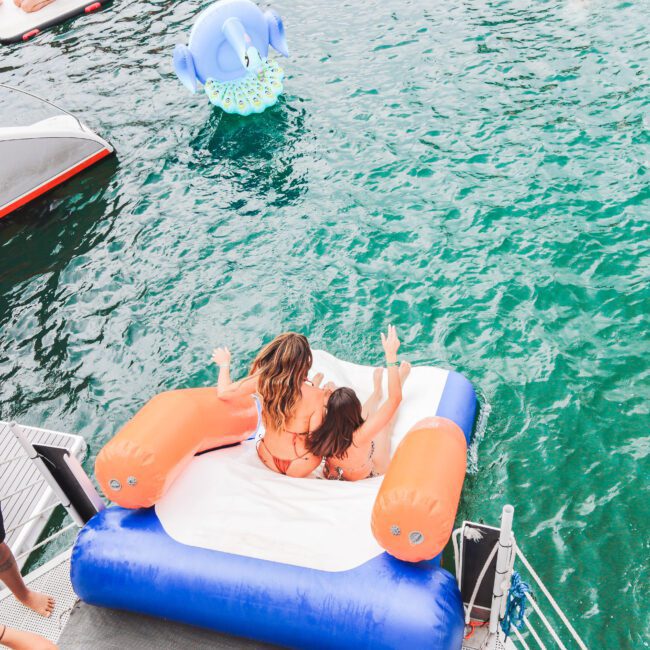 Two women sit on an inflatable float at the edge of a boat, preparing to slide into the water. A person in blue stands nearby, and a blue inflatable elephant floats in the water in the background.