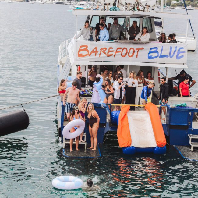 A group of people enjoy a boat party on the "Barefoot Blue," with some lounging, others swimming, and a few using an orange and white inflatable slide to enter the water. The scene is festive and lively.