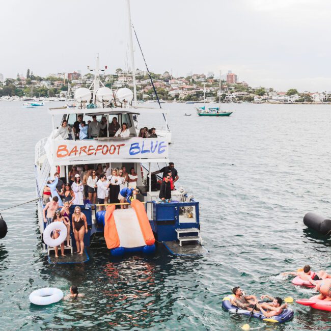 A lively group of people party on a boat called "Barefoot Blue" while others float nearby on inflatables in calm water; a cityscape is visible in the background under a cloudy sky.