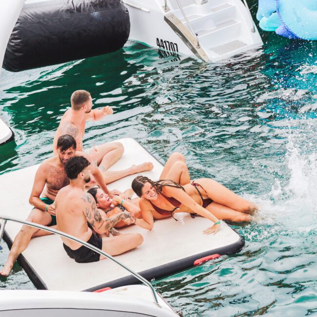 Five people in swimsuits relax and laugh on a floating mat in the water next to boats, as one person splashes into the water nearby. The scene is lively and playful on a sunny day.