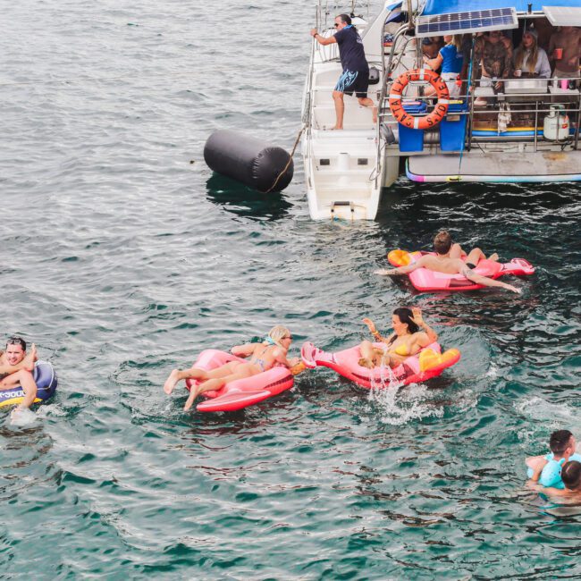 People relax on colorful pool floats and swim in the water near a boat with others on board. The scene is lively and joyful, suggesting a fun summer day on the water.