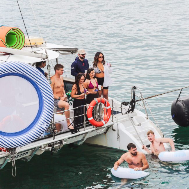 A group of people in swimwear enjoy a boat party on the water; some stand on the boat, while two men float nearby with inflatable tubes. Mats and a life ring are visible on the boat. The mood is relaxed and festive.
