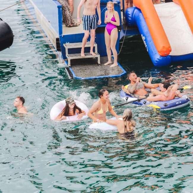 A group of people enjoy swimming and relaxing on inflatables in the water near a docked boat with an orange slide; some are climbing out of the water while others play and lounge.