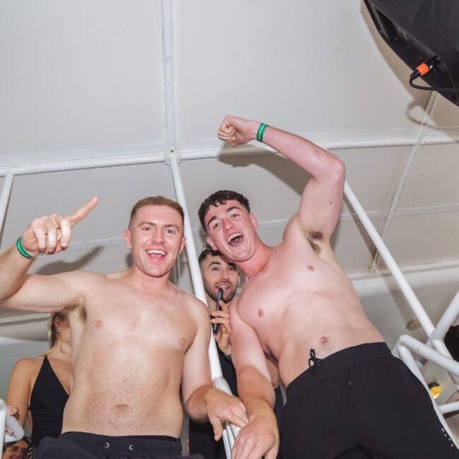 Two smiling young men, both shirtless and wearing black shorts, stand closely together on a white structure, celebrating with raised arms and excited expressions at what appears to be a lively indoor event or party.