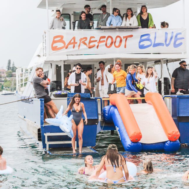 A group of people are enjoying a party on a boat named "Barefoot Blue," with some standing on the deck and others swimming or relaxing on inflatables in the water next to a blue and orange slide.