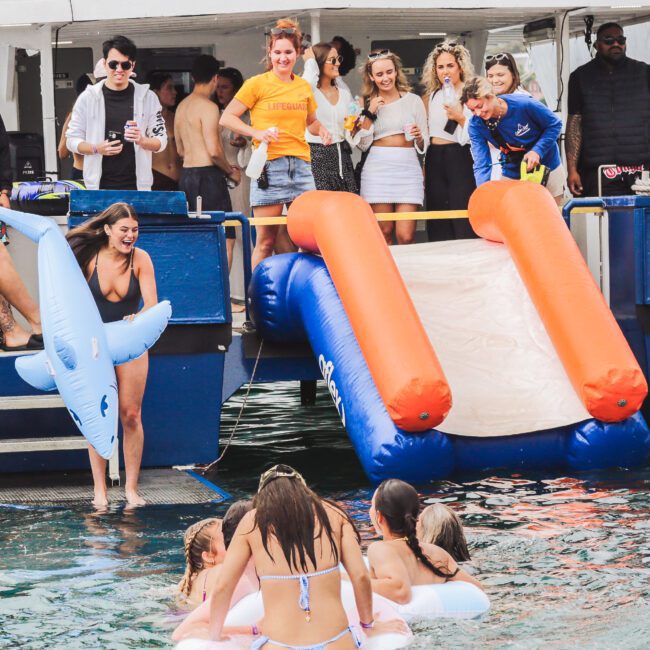 People enjoy a boat party with a water slide and inflatables. Several women in swimsuits are on a float in the water, while others on the boat watch and smile. A large sign reads "BAREFOOT BLUE.