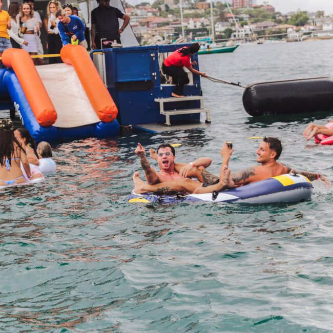 People swim and relax on floats near a docked boat with an orange and white slide. Some laugh and celebrate in the water while others sit on the boat, enjoying a sunny day by the shore.