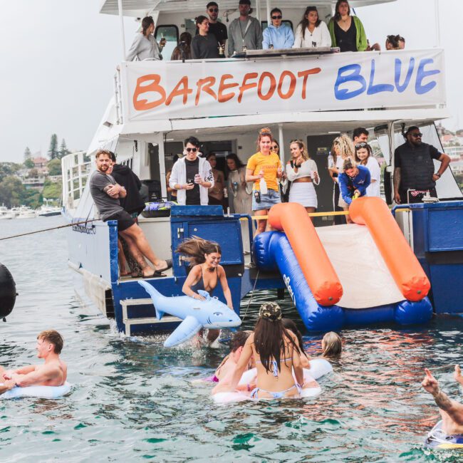 People enjoy a lively boat party on the water. Some are on inflatables or swimming, while others gather on the boat’s deck, which features a blue and orange slide. The boat's sign reads "Barefoot Blue.