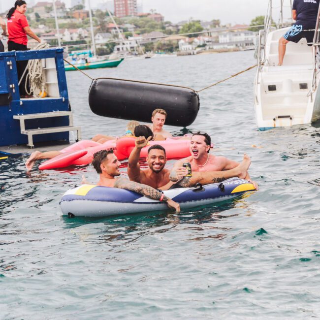 Three men smile and relax on an inflatable raft in the water near boats, while other people float on inflatables and a dock is visible in the background. The scene is cheerful and lively.