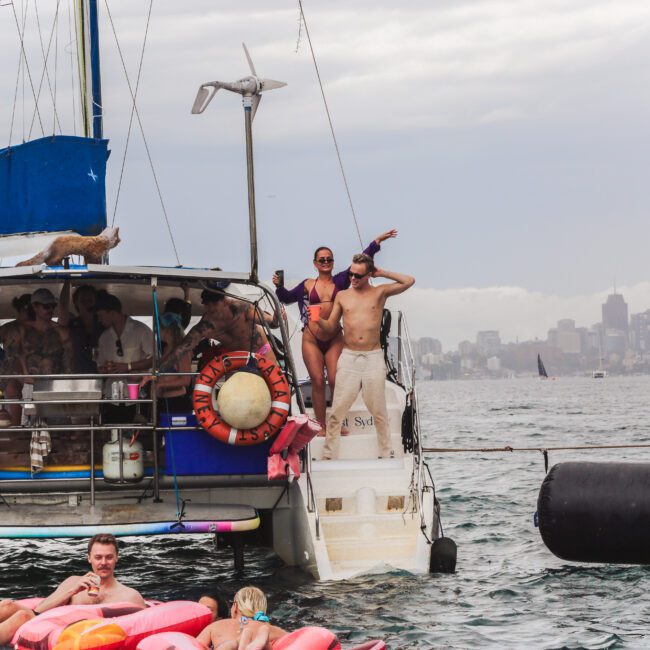 A group of people enjoy a party on a catamaran anchored in the water near a city skyline. Two people pose on the boat's swim platform, while others relax on inflatables in the water below. The sky is overcast.