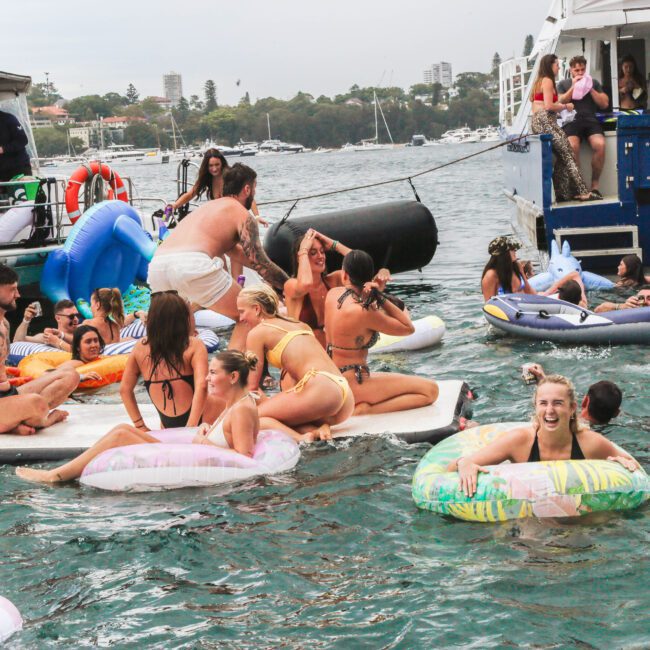 A group of people in swimsuits enjoy a lively party on the water, floating on inflatables and swimming near boats. Some people smile and laugh, while others relax on floaties in the busy, festive scene.