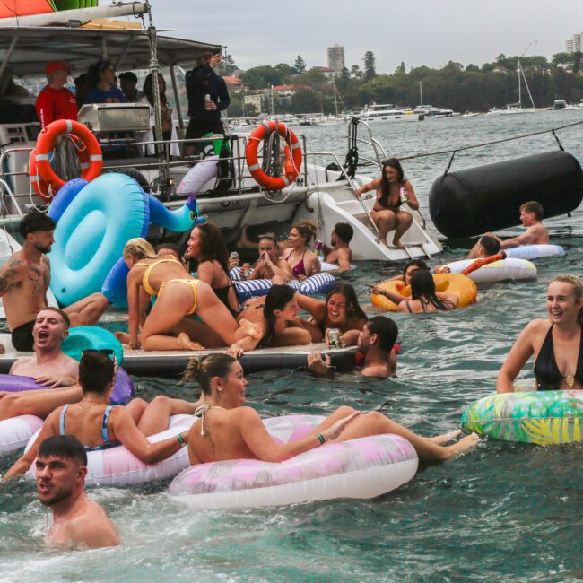 A group of people in swimsuits relax and have fun on colorful inflatable tubes in the water near boats, with others socializing on the boats. The scene looks lively, festive, and takes place on a cloudy day.