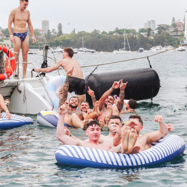 A group of young adults in swimsuits enjoy themselves on inflatable rafts in the water near a boat, smiling and making peace signs, with trees and buildings visible on the shore in the background.