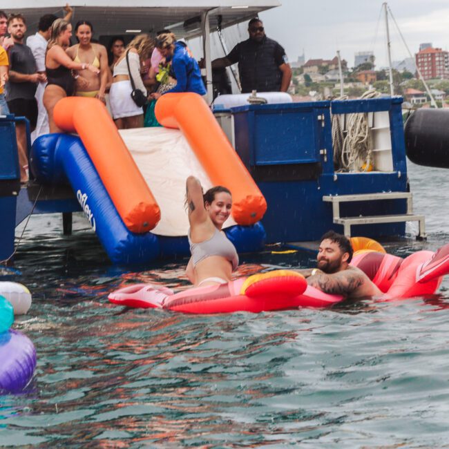 People relax and have fun on colorful floaties in the water near a boat with an inflatable slide. Some are in swimwear, smiling and waving, while others socialize on the boat. The background shows a cityscape and cloudy sky.