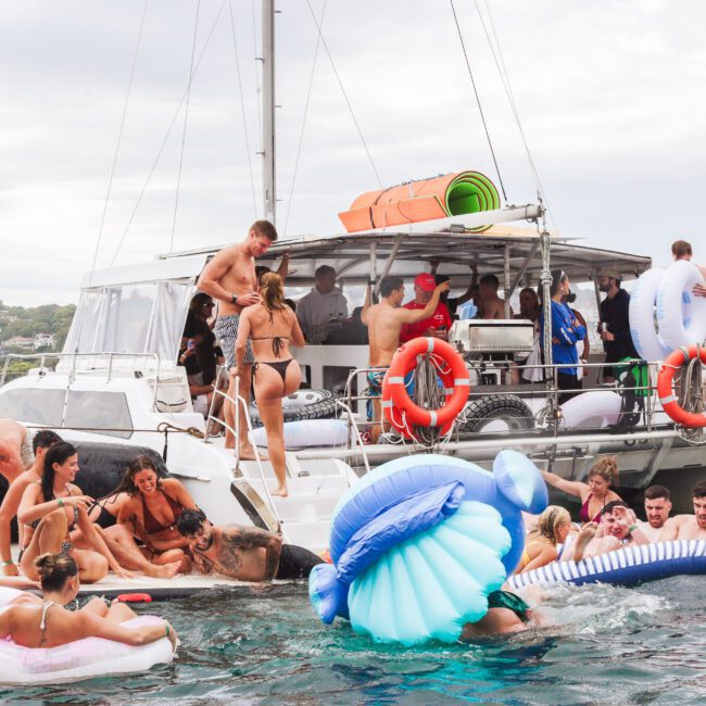 A group of people in swimsuits enjoy a party on and around a boat, with some on inflatables in the water and others on the boat. The scene is lively, with laughter and interaction under a cloudy sky.