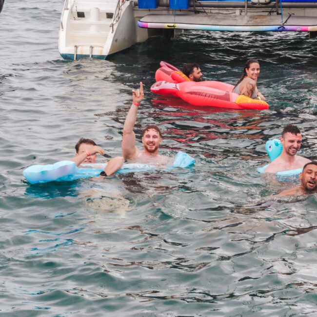 A group of people relax on colorful floaties in the water near a boat, smiling and waving at the camera. Some float on their backs, while others paddle or swim nearby. The scene is lively and joyful.