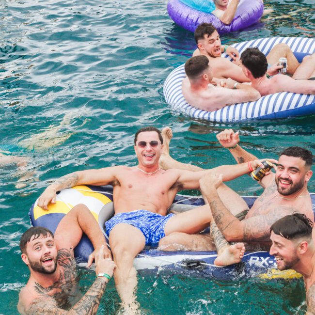 A group of smiling young men with tattoos relax on colorful pool floats in clear blue water, laughing and enjoying themselves in a lively, fun atmosphere.