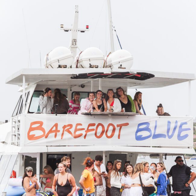 A group of people stand and sit on the upper and lower decks of a white boat with a large "Barefoot Blue" banner, docked on a calm body of water with houses and other boats visible in the background.