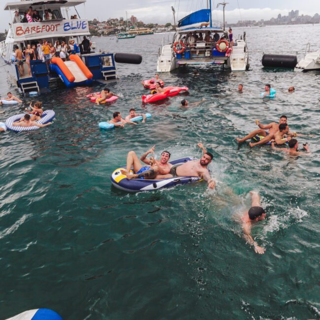 People relax on inflatable floats and swim in the ocean near anchored boats, enjoying a lively party atmosphere. Some sit on the boats, while others float or swim in the water under a cloudy sky.
