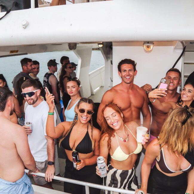 A group of people in swimwear enjoy a lively boat party, smiling, holding drinks, and taking selfies. The scene is bright and festive, with water and part of the boat visible in the background.