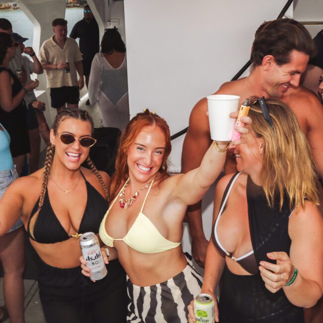 A group of people in swimwear laugh and pose with drinks at a lively boat party, with the ocean in the background. Several women in the foreground smile and raise their cans and cups toward the camera.