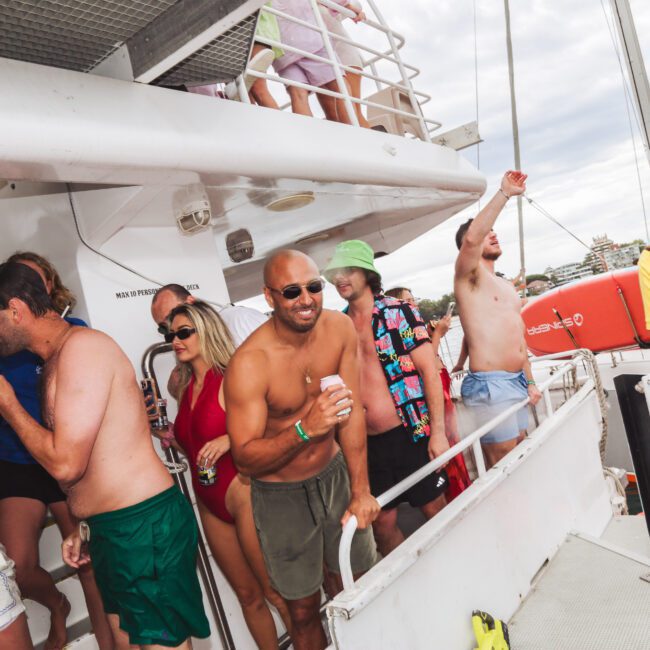 A group of people in swimwear enjoy a lively party on a boat. Some are dancing, others are talking and holding drinks. A woman in a yellow shirt stands by the railing while the mood is festive and relaxed.