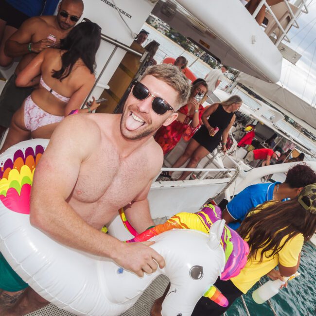 A man wearing sunglasses and a unicorn float smiles and sticks out his tongue on a crowded boat with people enjoying a party near the water.